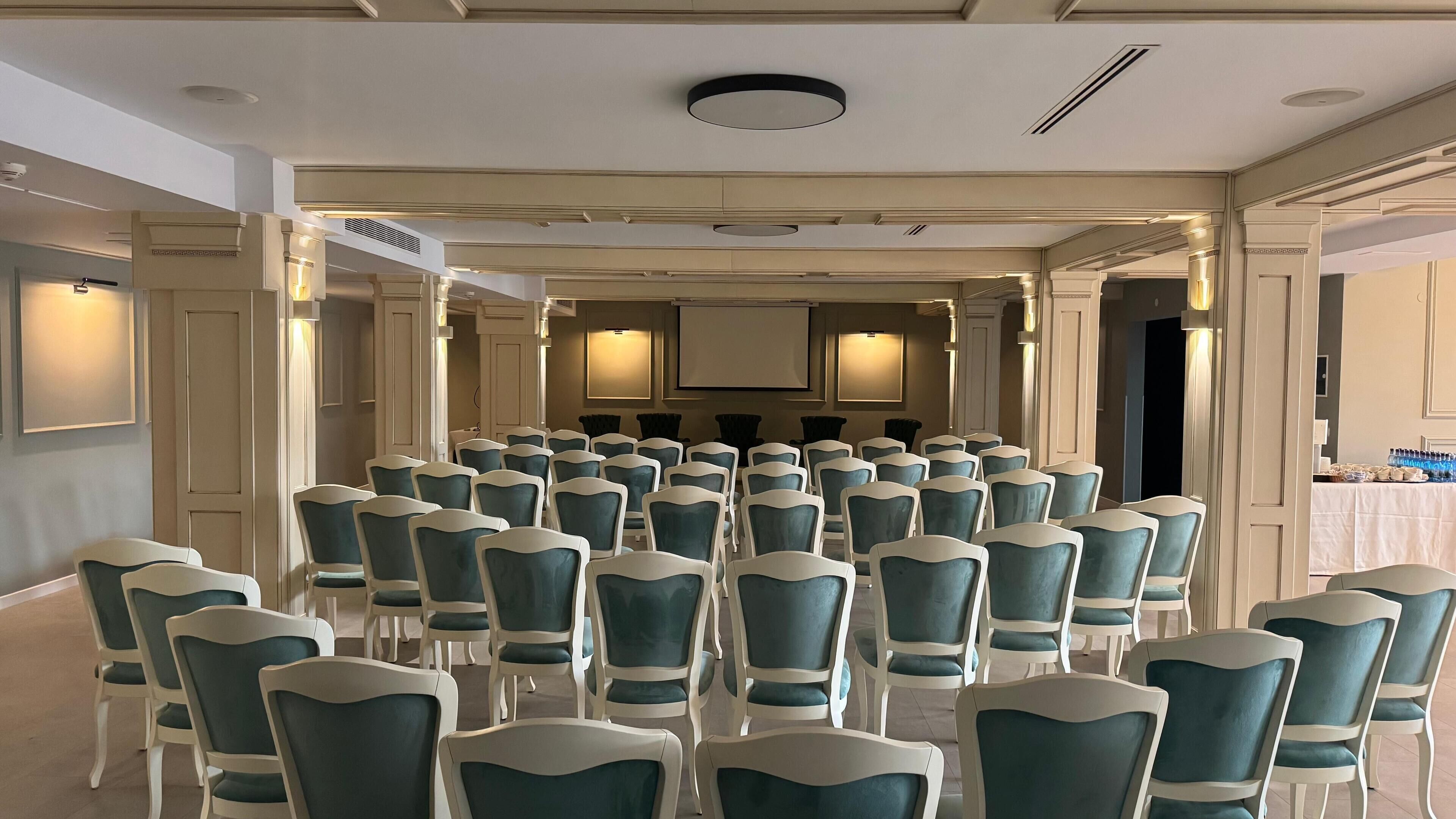 A conference room with rows of teal chairs and white frames, a large screen at the front, and recessed lighting.