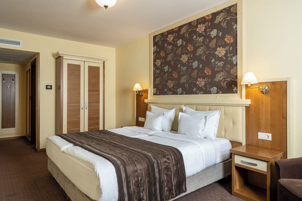 A hotel room with a large bed, two bedside tables, a nightstand, a wardrobe, and a floral wallpaper.
