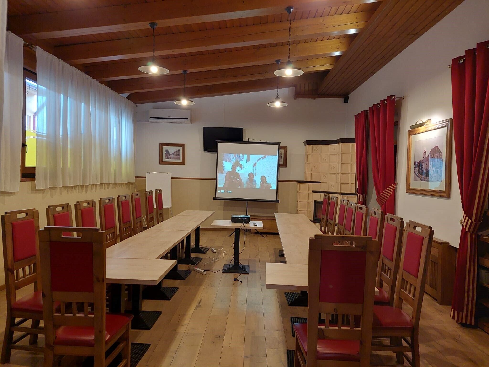 Un salón de conferencias con mesas de madera y sillas rojas, una pantalla de proyector y un televisor en la pared.
