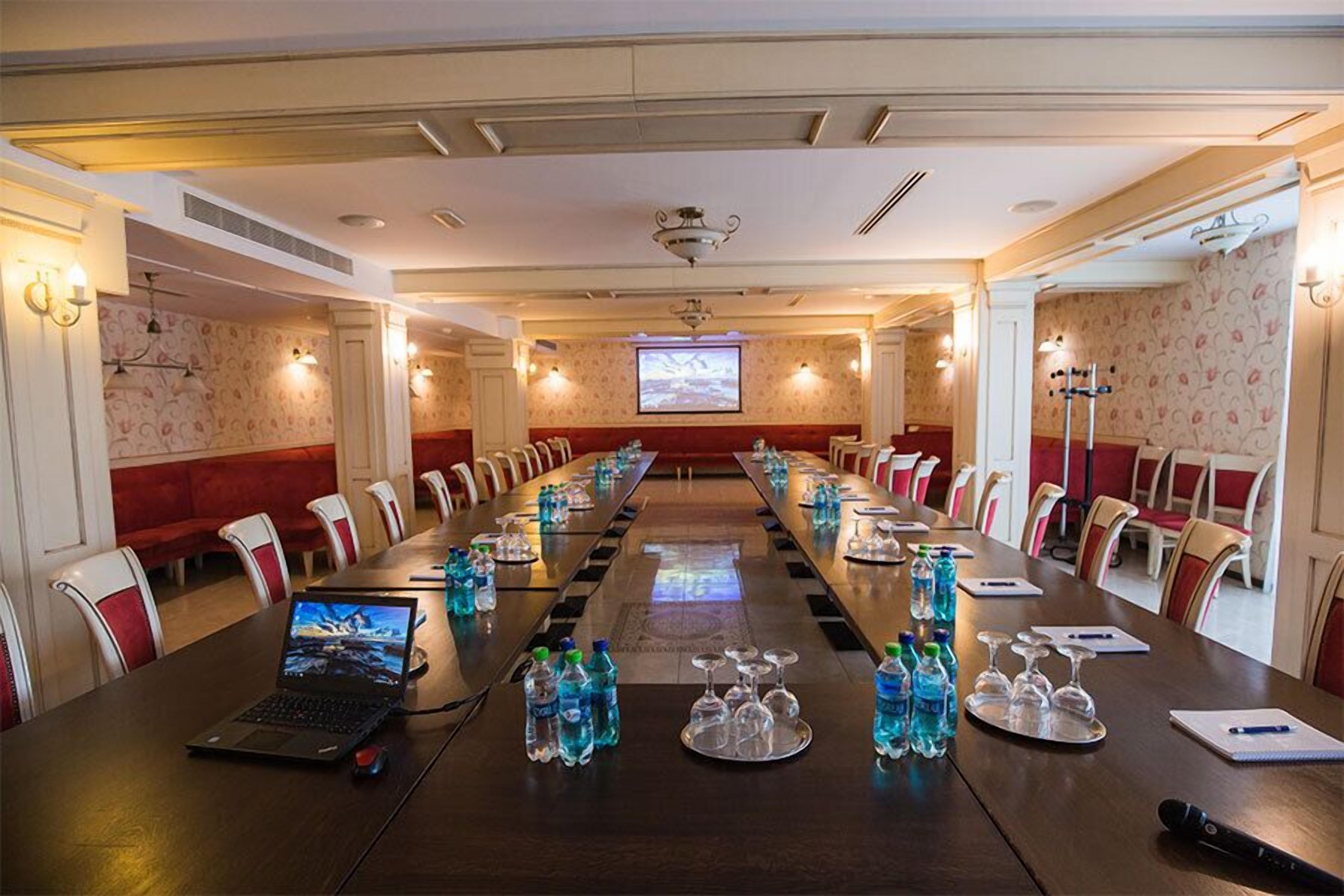 Un salón de conferencias con una larga mesa, sillas, portátiles, botellas de agua y una pantalla de proyector.
