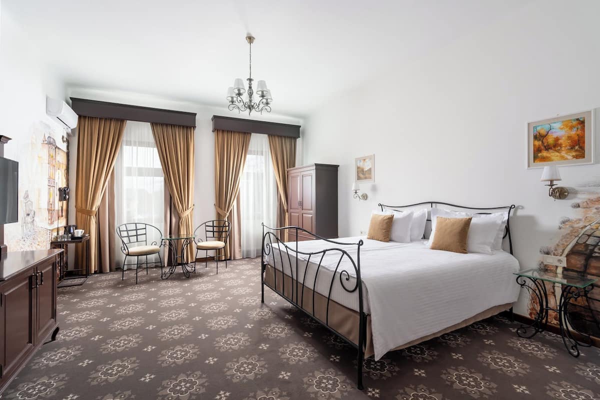 A hotel room with a bed, a desk, two chairs, a wardrobe, and a painting on the wall.