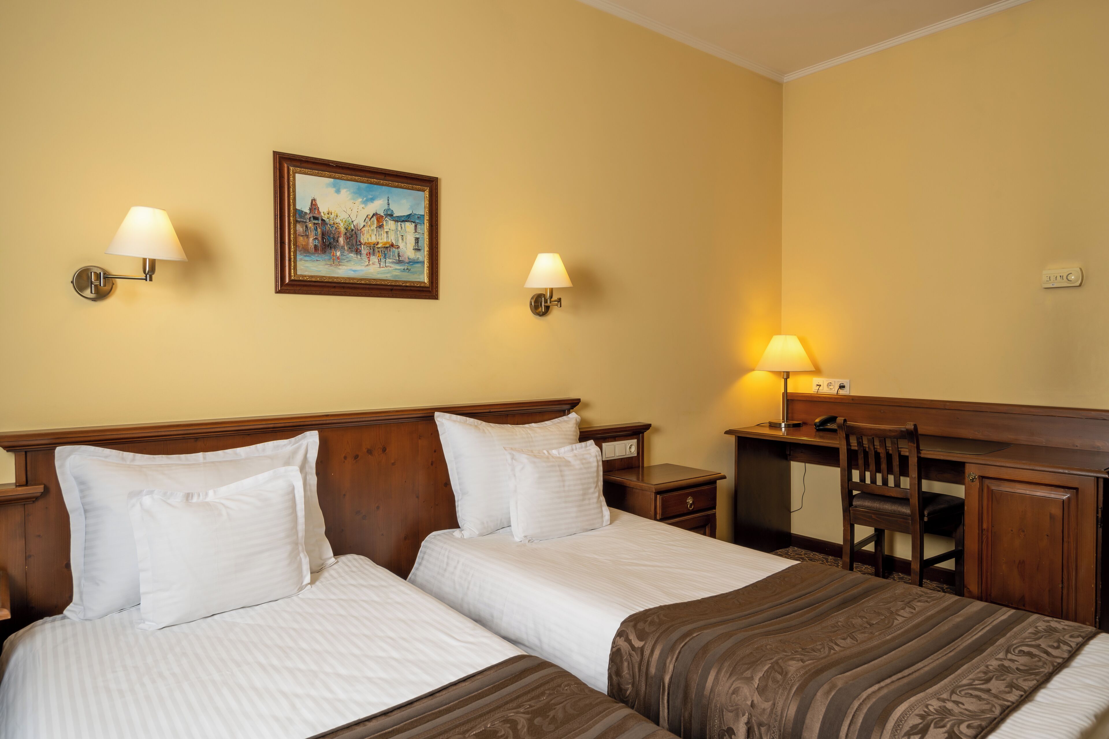 A hotel room with two beds, a desk, a chair, and a painting on the wall.