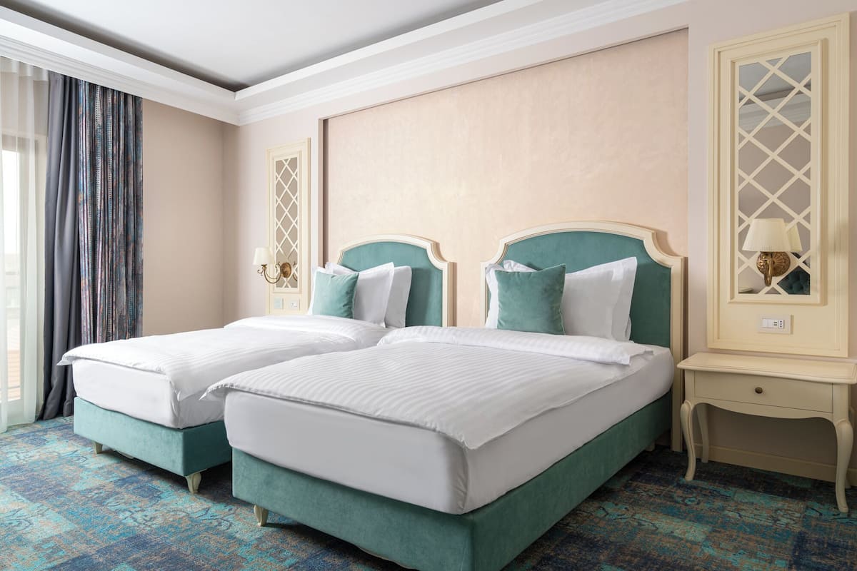 A hotel room with two single beds, a small bedside table, a mirror, and a window with curtains.
