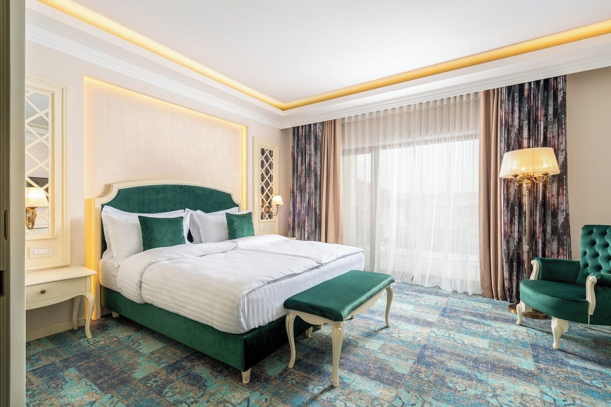 A hotel room with a large bed, a bench, a green armchair, and a patterned carpet.
