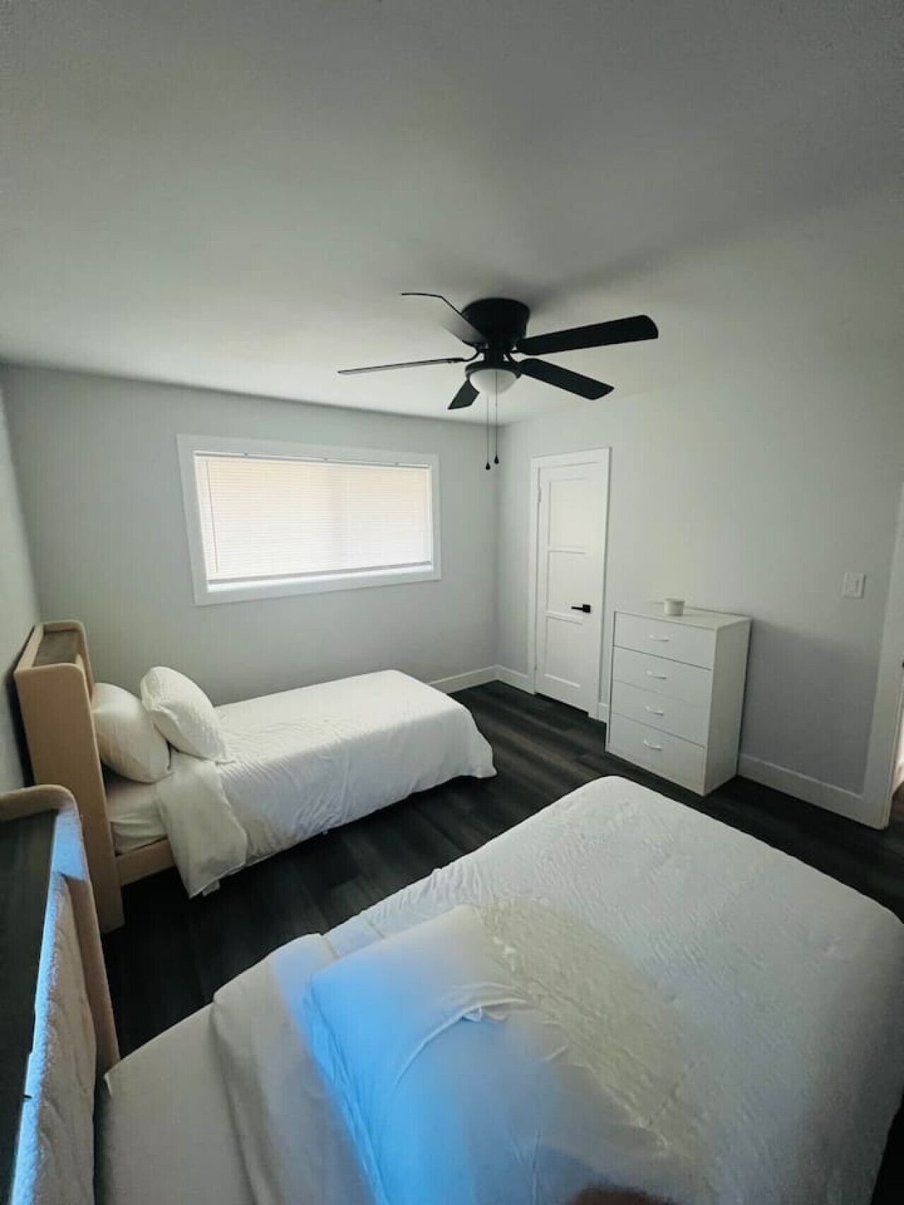 Une chambre avec deux lits simples, un ventilateur de plafond, une fenêtre avec stores, une armoire blanche et une commode.
