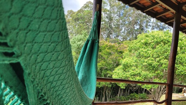 A hammock hanging under a wooden structure with a tiled floor and a view of a grassy area and trees.