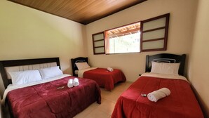 A hotel room with three beds, each with red bedding and white pillows. There is a window with wooden frames and a view of greenery outside.