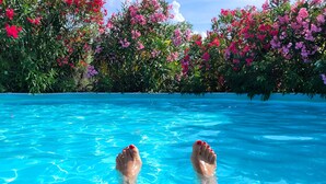 Une piscine à l’eau d’un bleu cristallin, avec des ongles de pieds peints en rouge, entourée d’une végétation luxuriante aux fleurs roses et violettes.