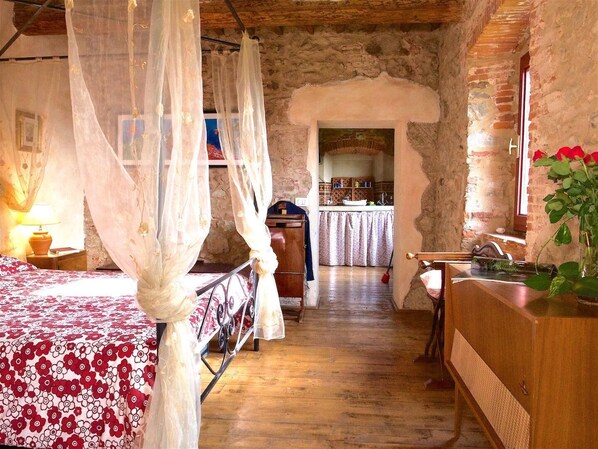 A rustic room with a bed under a canopy, a wooden dresser, and a kitchen area visible through an archway. - Agriturismo I Pitti (Serravalle Pistoiese)