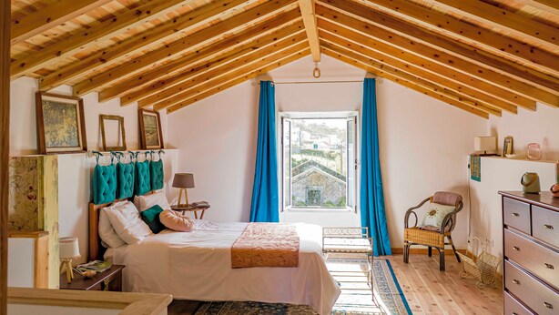 A bedroom with a sloped wooden ceiling, a bed with white bedding, a window with blue curtains, and a view of buildings outside. - Candelaria House (Candelária)