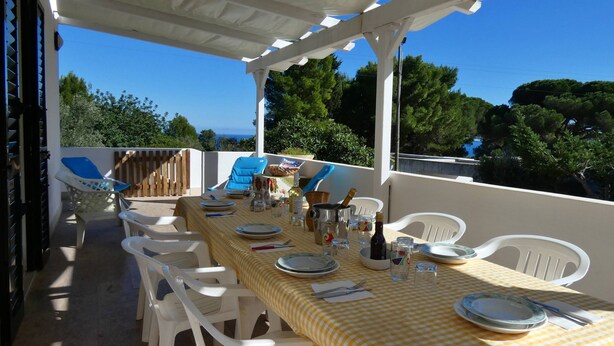 An outdoor dining area with a checkered tablecloth, white plastic chairs, and a view of the sea.
