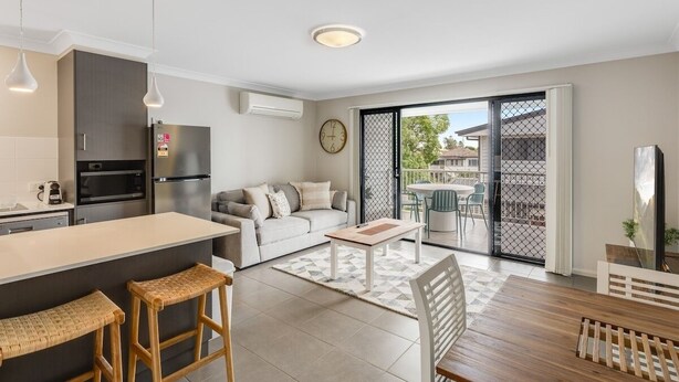 Une cuisine moderne dotée d’un îlot central, un coin repas avec une table et des chaises en bois, un canapé et une porte-fenêtre coulissante donnant sur un balcon.