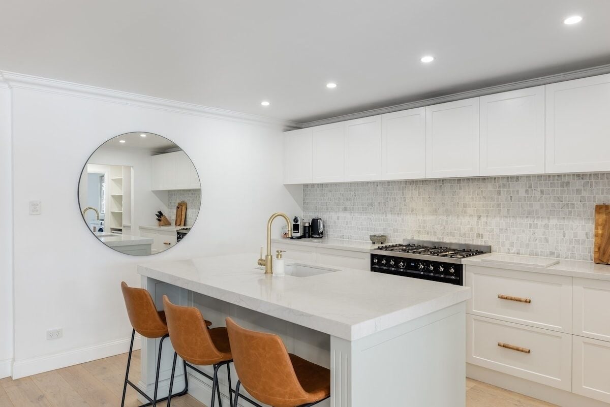 A modern kitchen with white cabinetry, a large island with bar stools, a round mirror, and a tiled backsplash.