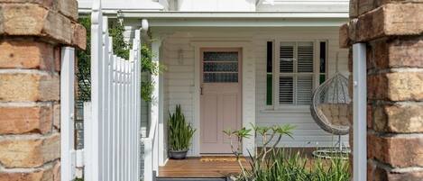 Une maison blanche avec un porche en bois, une porte rose et une clôture blanche.