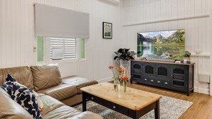 Una sala de estar con un sofá de cuero, una mesa de centro de madera con un jarrón de flores, un mueble de entretenimiento negro y un televisor de pantalla plana grande que muestra un paisaje de montaña.