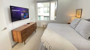 Ein Schlafzimmer mit großem Flachbildfernseher an der Wand, einer hölzernen Kommode, einem Bett mit weißer Bettwäsche, einem kleinen Hocker und einem Fenster mit Blick ins Freie.