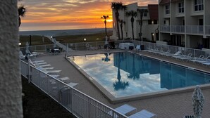 Ein Swimmingpool mit Liegestühlen und Sonnenschirmen, umgeben von einem Zaun, mit einem Sonnenuntergang im Hintergrund.