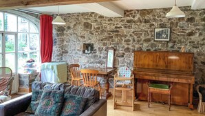 A cozy living room with a stone wall, a piano, a sofa with patterned cushions, and a dining area with wooden chairs.