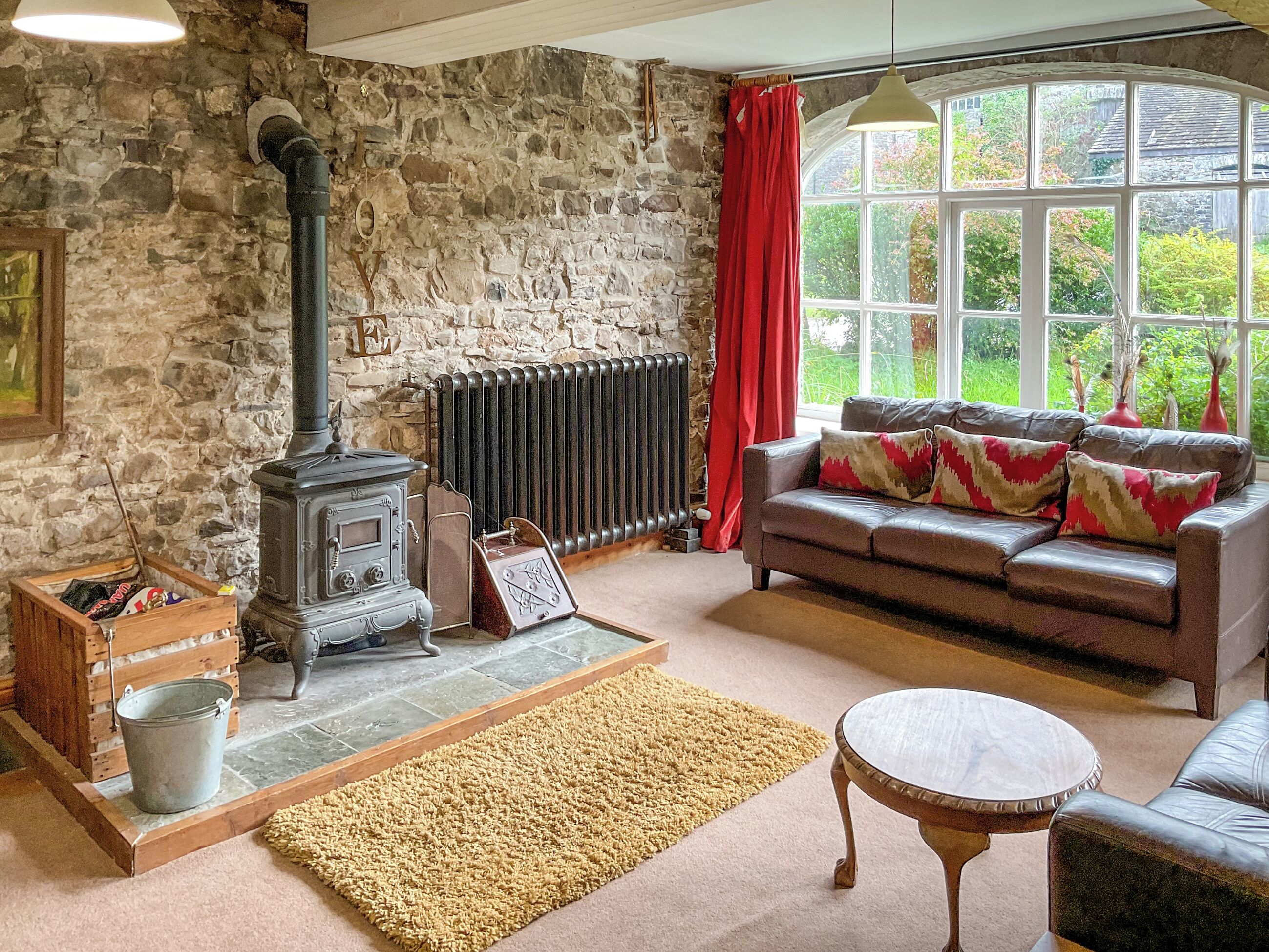 A cozy living room with a stone wall, a vintage stove, a leather sofa, and a large window with a view of greenery.