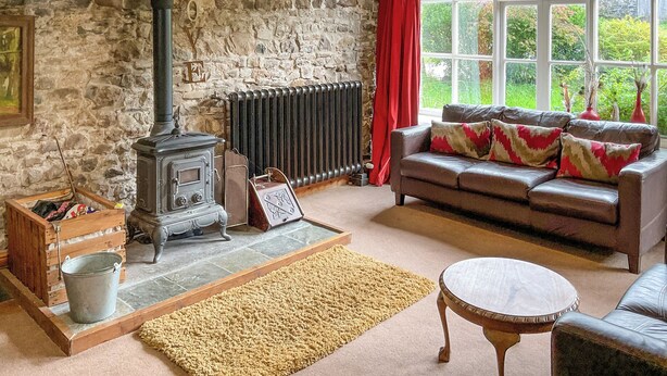 A cozy living room with a stone wall, a vintage stove, a leather sofa, and a large window with a view of greenery.