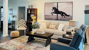 Un salon spacieux avec un ventilateur de plafond, un grand tableau représentant un cheval, un canapé, un fauteuil bleu et une table basse avec des livres et un vase.