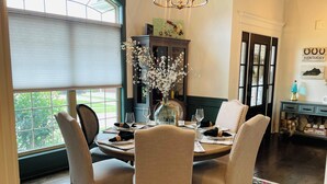 Un coin repas avec une table ronde dressée pour quatre personnes, entourée de chaises rembourrées. Un vase avec des fleurs blanches est posé sur la table, et on aperçoit en arrière-plan un buffet en bois surmonté d’un élément décoratif.