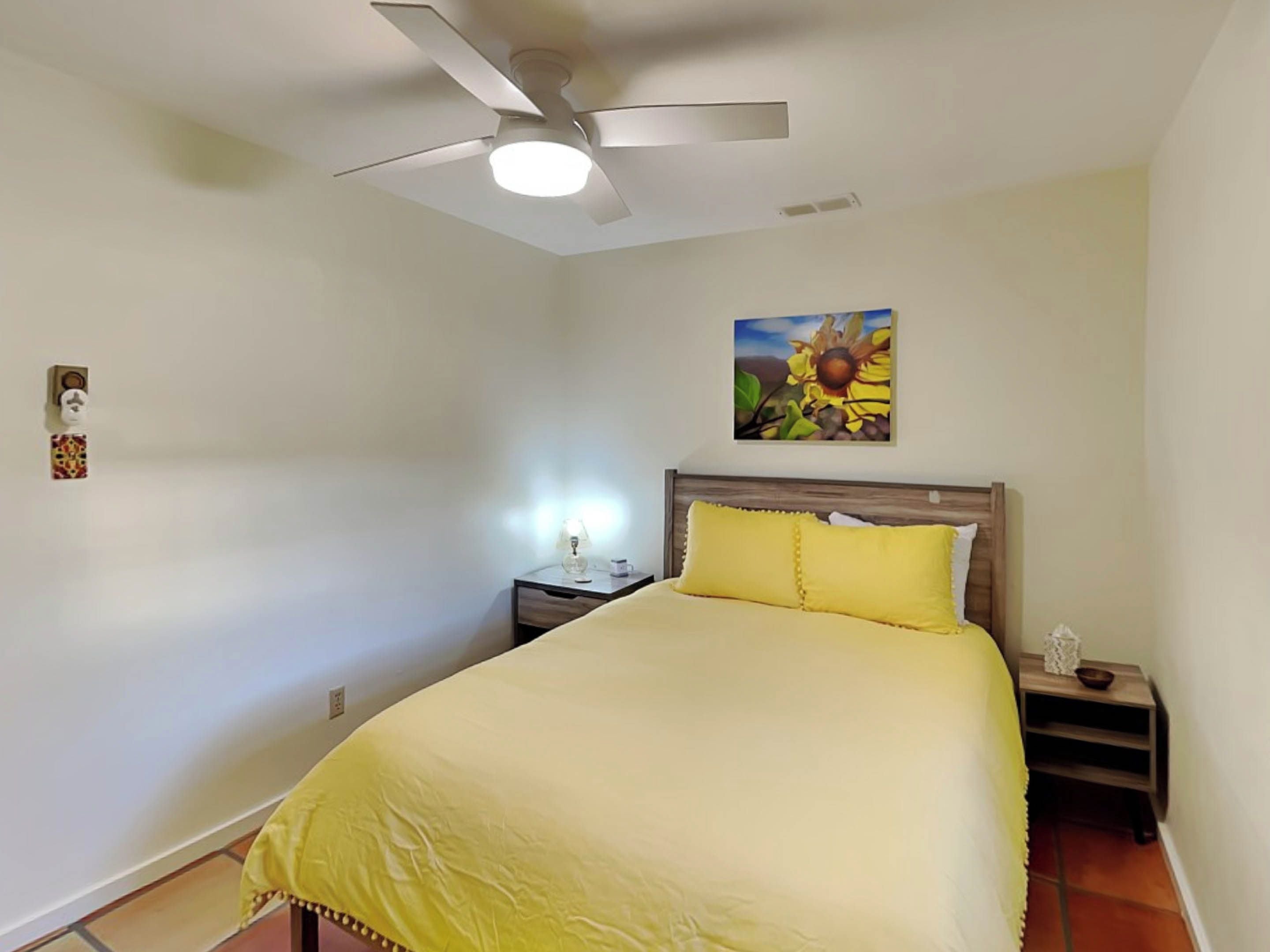Un dormitorio con cama, mesita de noche, lámpara, ventilador de techo y un cuadro de girasoles.