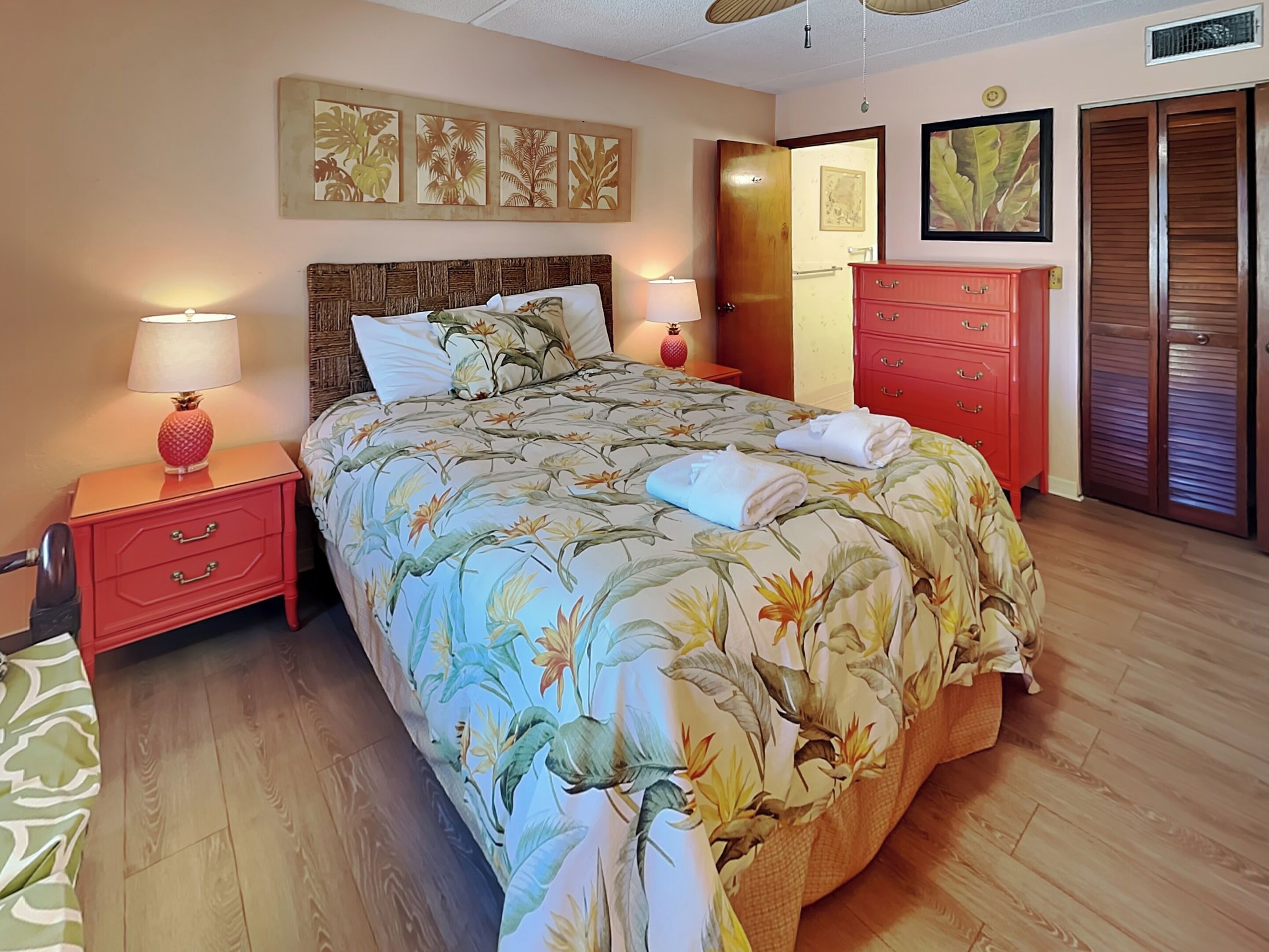 A neatly made bed with floral bedding, a red nightstand with a lamp, and a red dresser with a mirror.