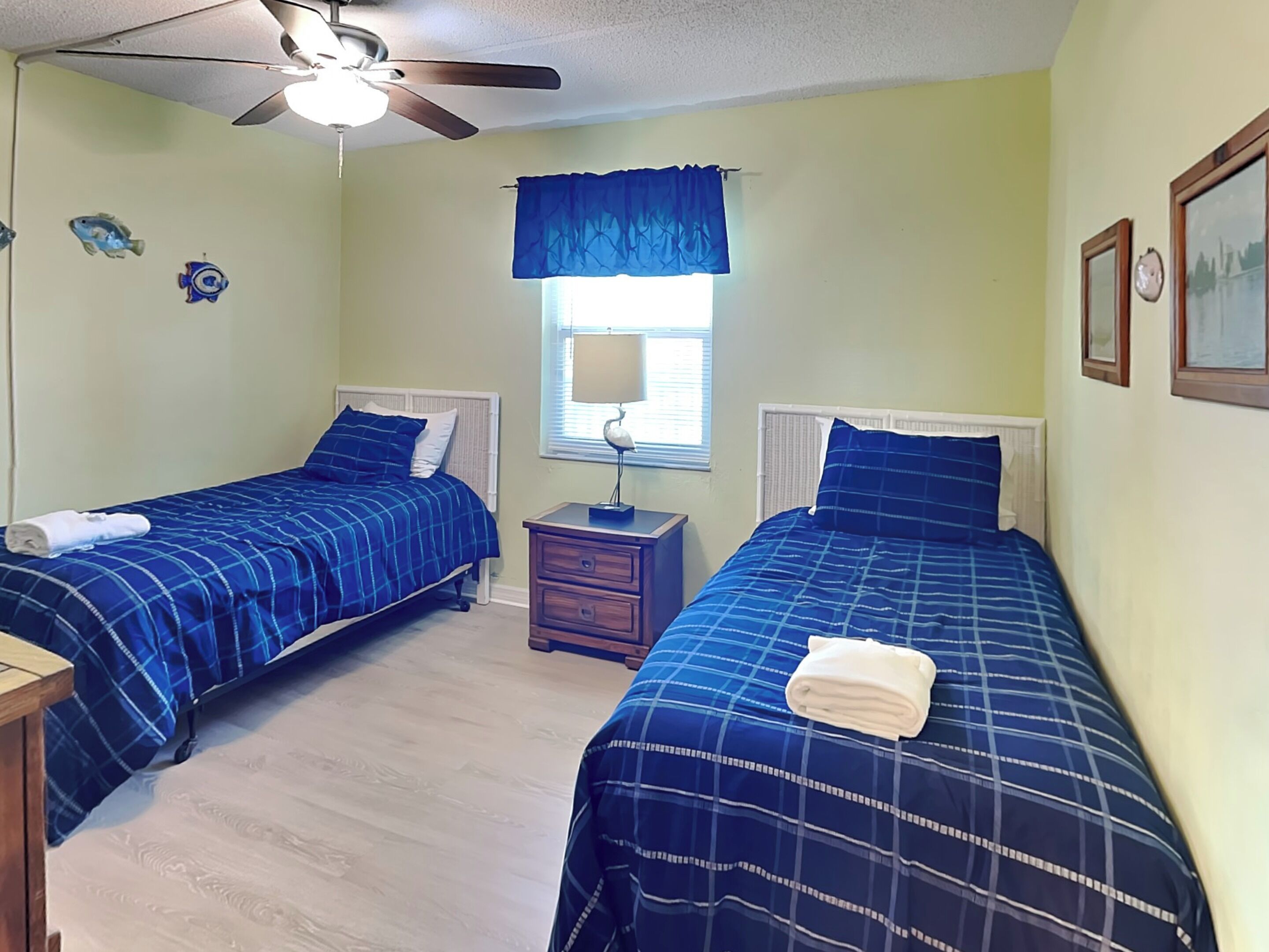 A bedroom with two beds, a ceiling fan, a window with a blue curtain, and a small table with a lamp.