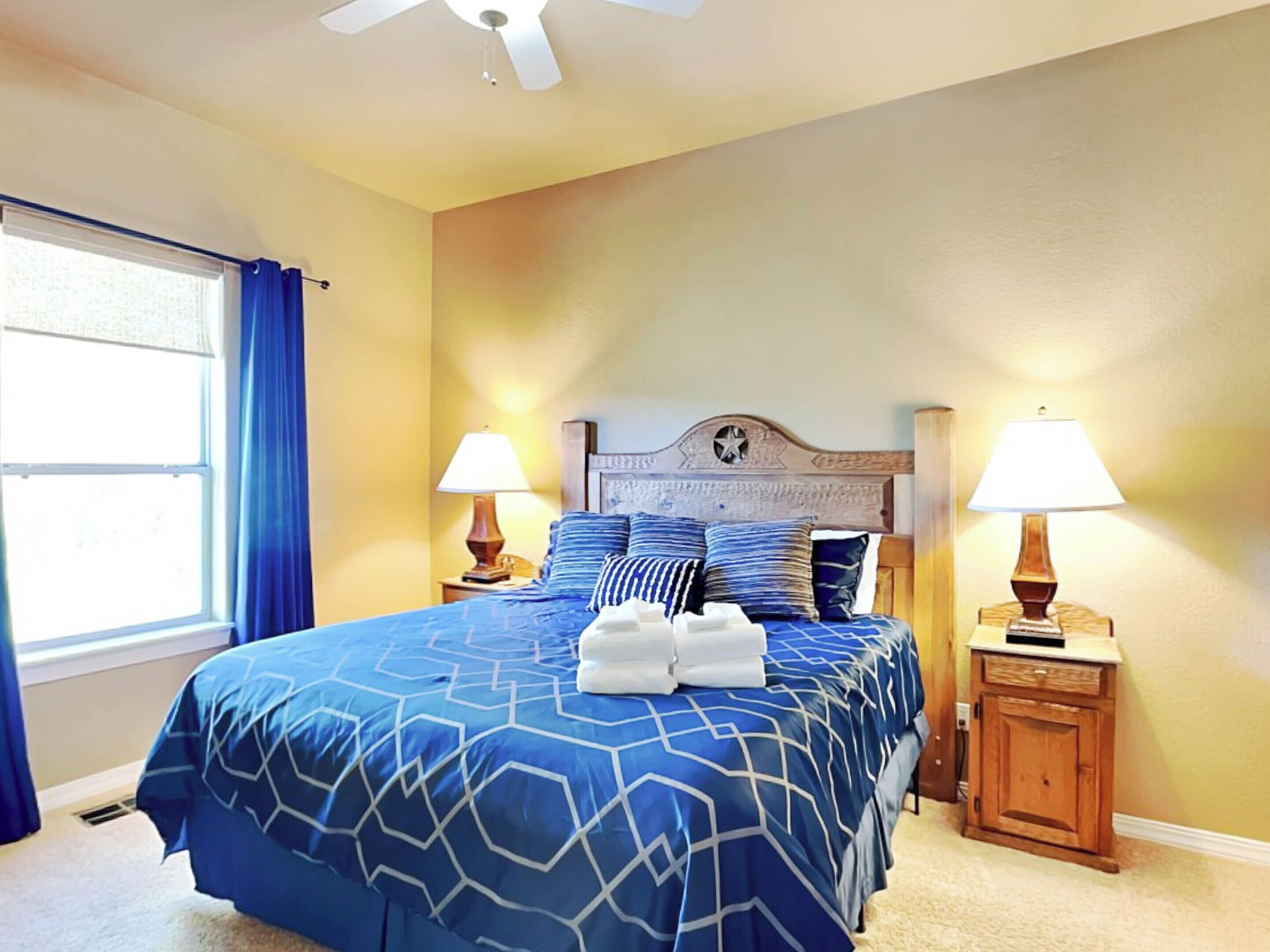 A bedroom with a large bed, two bedside tables with lamps, a ceiling fan, and a window with curtains.