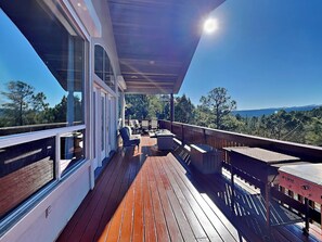 Una terraza de madera con mobiliario exterior, un espacio cubierto y una vista panorámica de árboles y montañas.