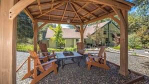 A wooden gazebo with a table and four chairs, surrounded by gravel and greenery. - Cody's Comfy Cabin-Near Yosemite (Mariposa)