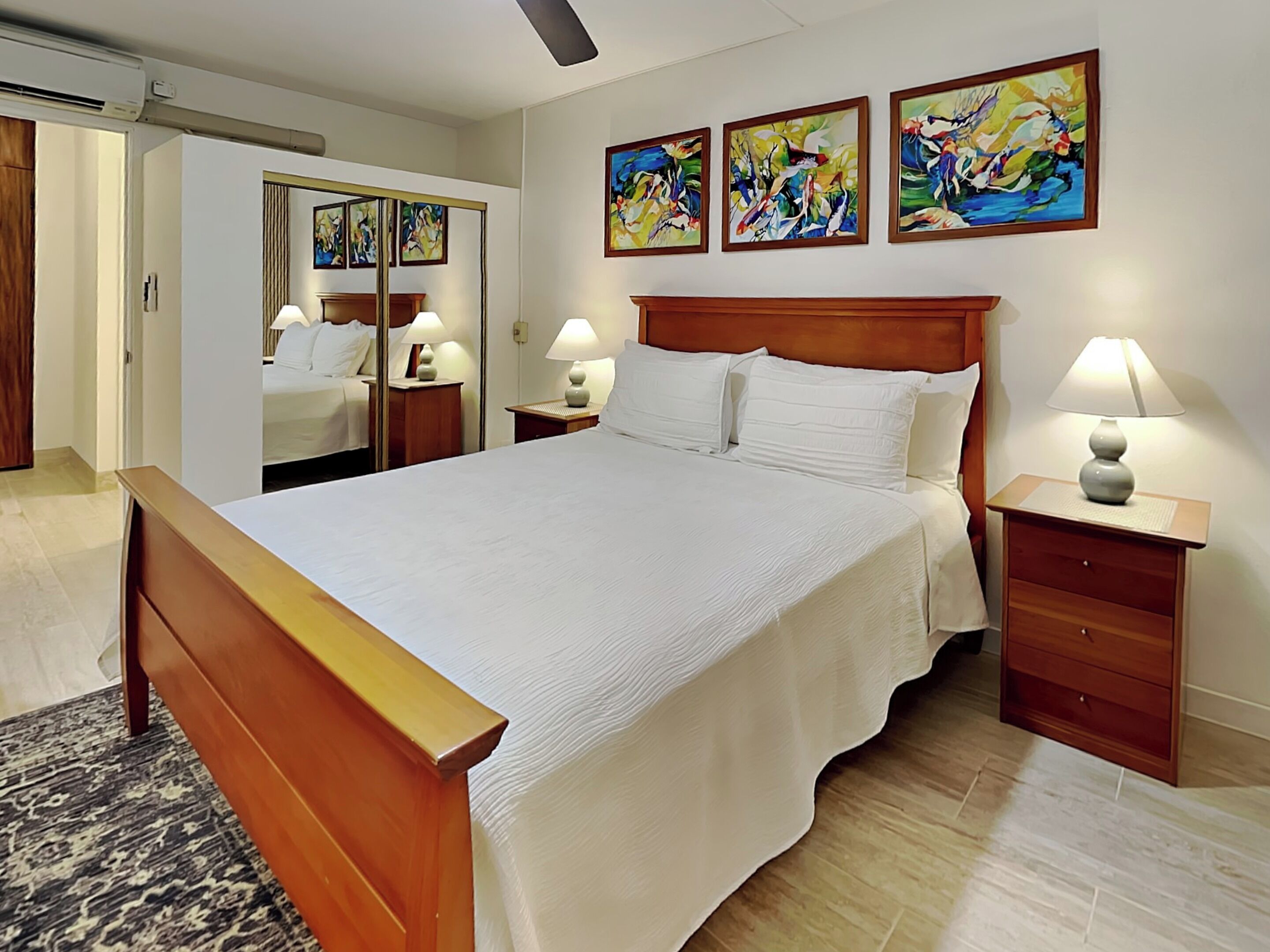 A hotel room with a large bed, wooden headboard, two bedside tables with lamps, a mirror, and three abstract paintings on the wall.