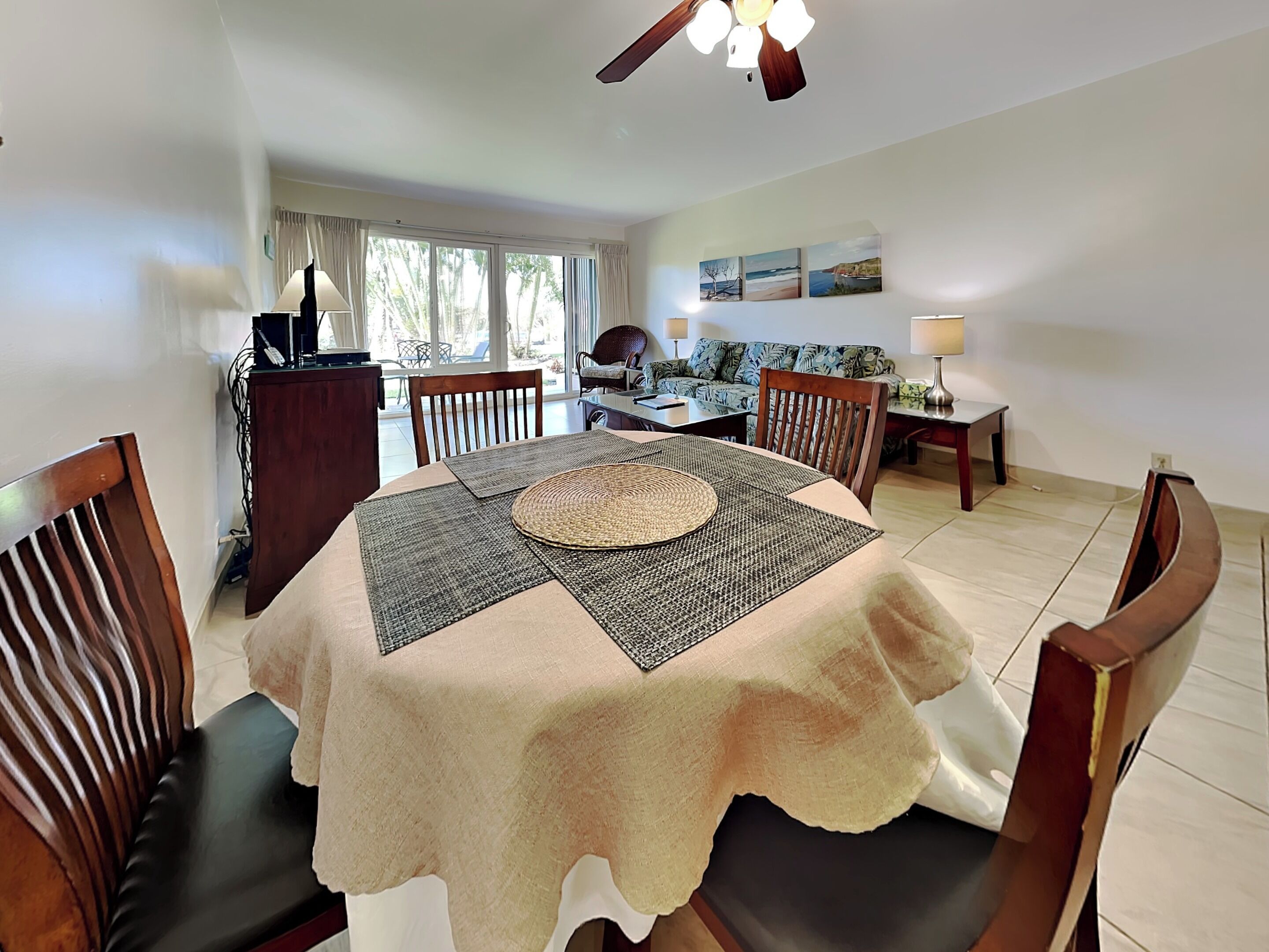 A dining area with a round table, chairs, a ceiling fan, and a view of the outdoors through large windows.