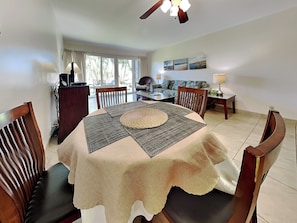A dining area with a round table, chairs, a ceiling fan, and a view of the outdoors through large windows. - Kihei Akahi D-105 by Maui Rental Group (Kihei)