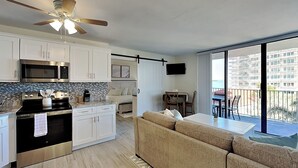 A modern kitchen with white cabinets, a microwave, and a stove. There is a beige sofa and a dining table with chairs. A ceiling fan is visible, and the room has a large window. - Lovers Key Beach Club by Distinctive Beach Rentals (Fort Myers Beach)