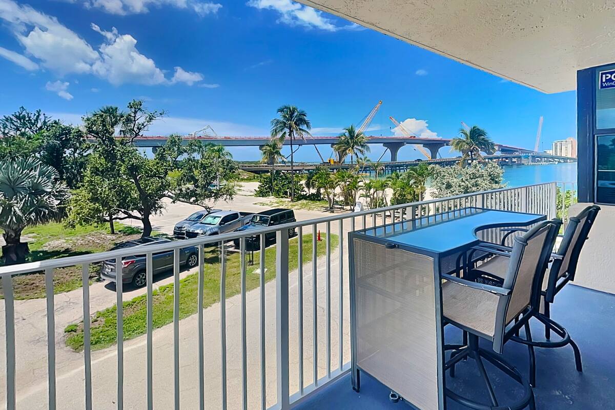 Ein Balkon mit Tisch und Stühlen, der auf eine Straße mit parkenden Autos und eine Brücke im Hintergrund blickt.
