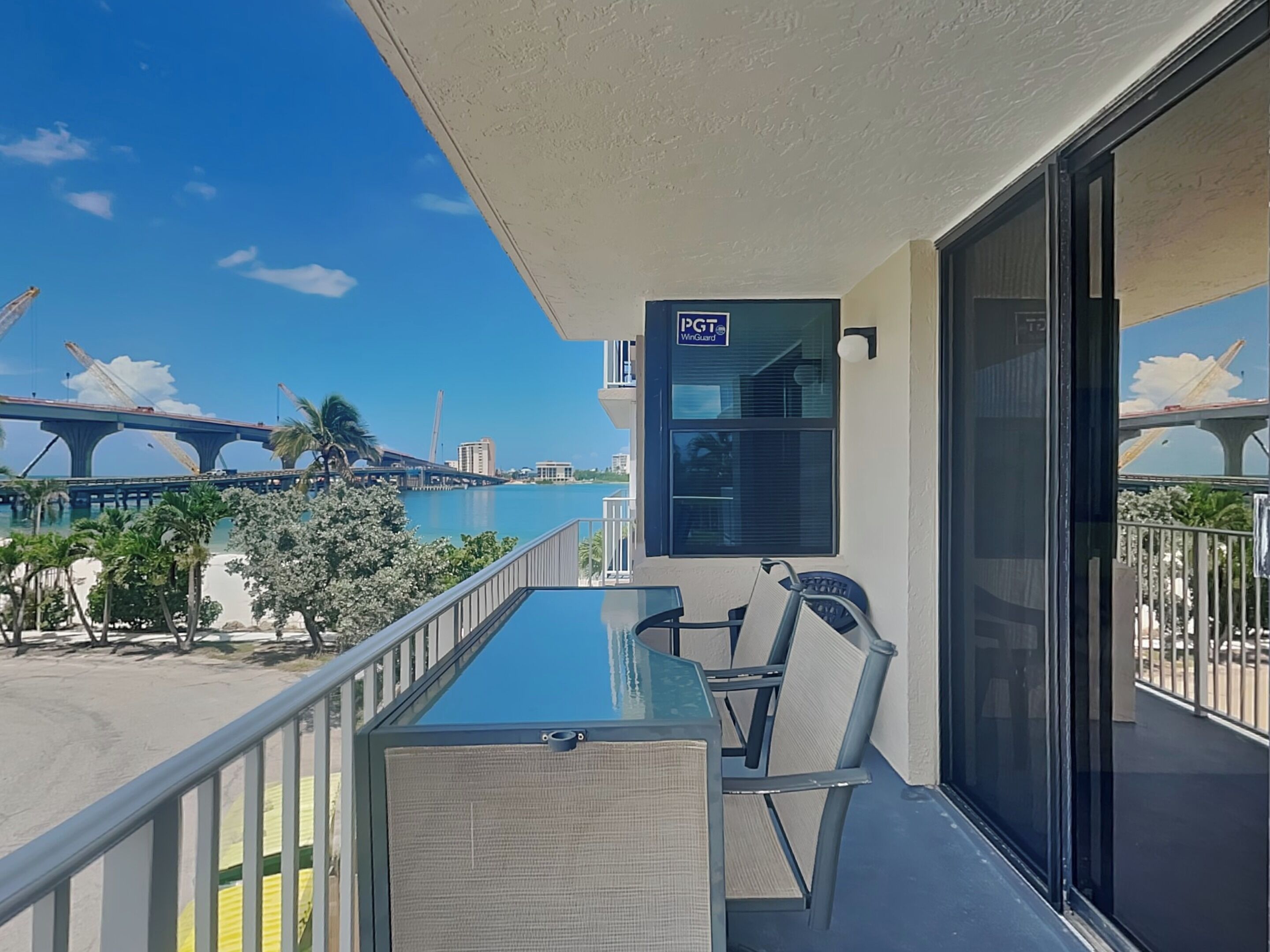 Un balcon avec une table et des chaises en verre, donnant sur une plage et un pont.