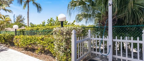 A gated entrance with a white picket fence, a metal gate, and a concrete pathway leading to a palm tree and a blue sky.