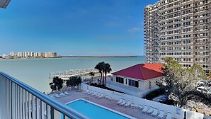 A pool area with lounge chairs, a small building with a red roof, and a view of a body of water and distant buildings. - Lovers Key Beach Club by Distinctive Beach Rentals (Fort Myers Beach)