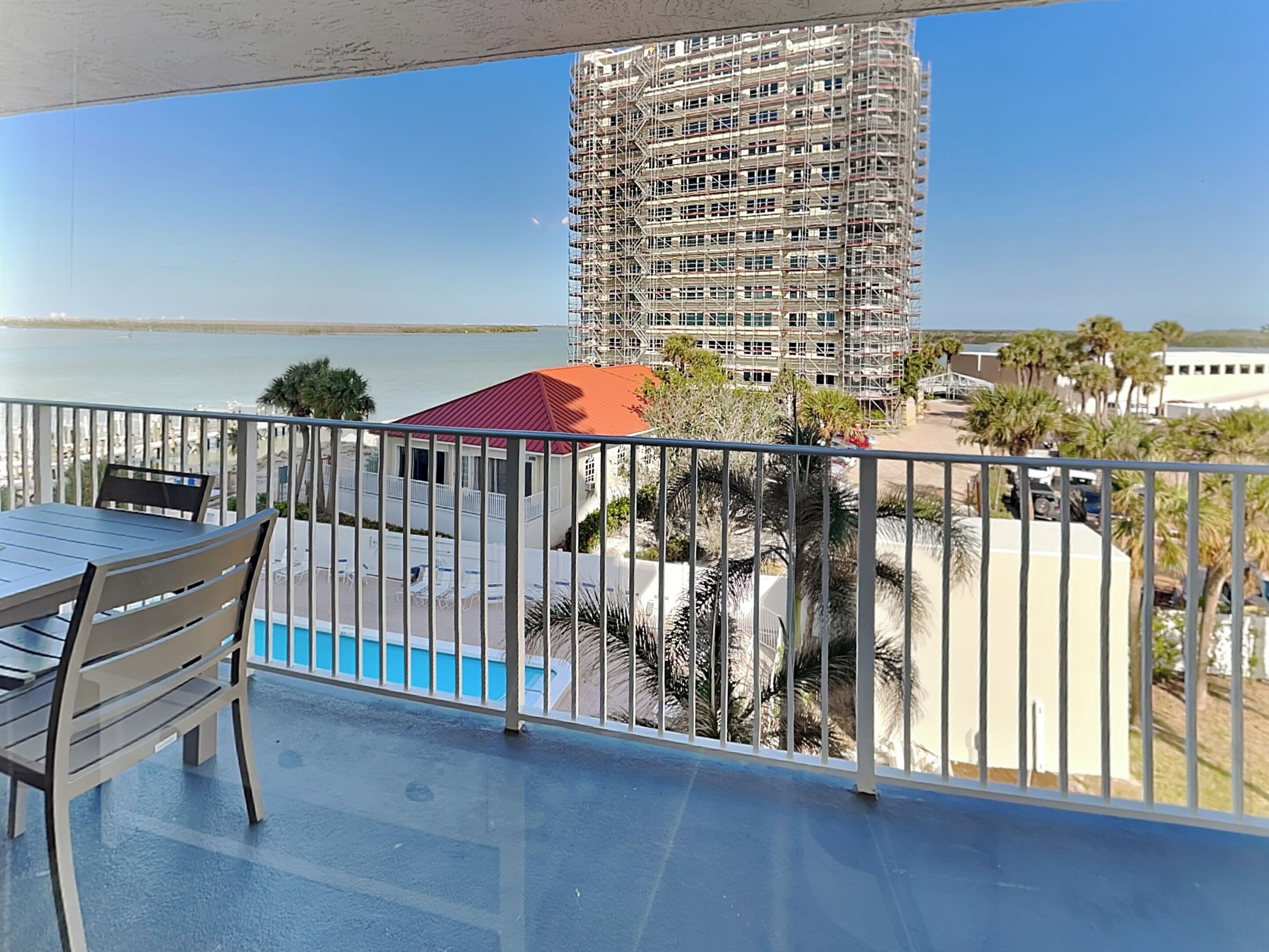 A balcony with a view of a building under construction, a pool, and palm trees.