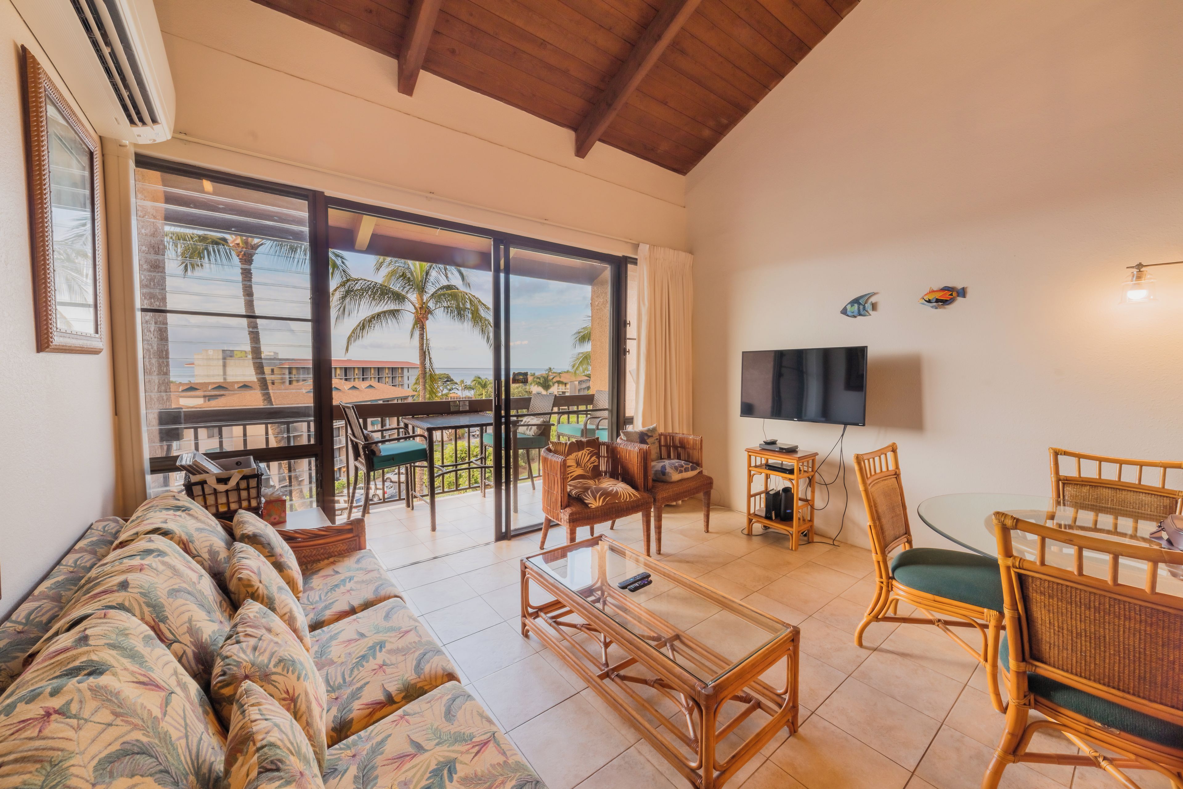 A spacious living area with a sofa, coffee table, and dining set. A large sliding glass door leads to a balcony with a view of palm trees and buildings.