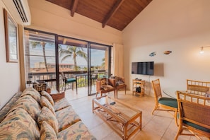 A spacious living area with a sofa, coffee table, and dining set. A large sliding glass door leads to a balcony with a view of palm trees and buildings. - Maui Vista 3402 by Maui Rental Group (Kihei)