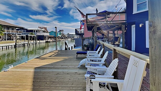 A wooden pier with white lounge chairs and a small table, overlooking a body of water with buildings in the background. - Siesta Keys by Miss Kitty’s (Rockport)