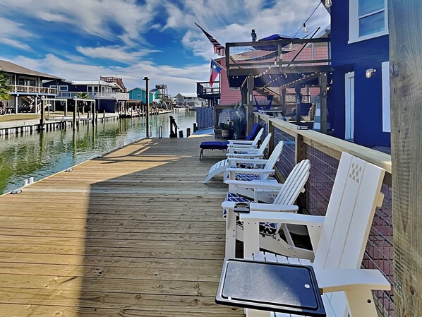 A wooden pier with white lounge chairs and a small table, overlooking a body of water with buildings in the background.