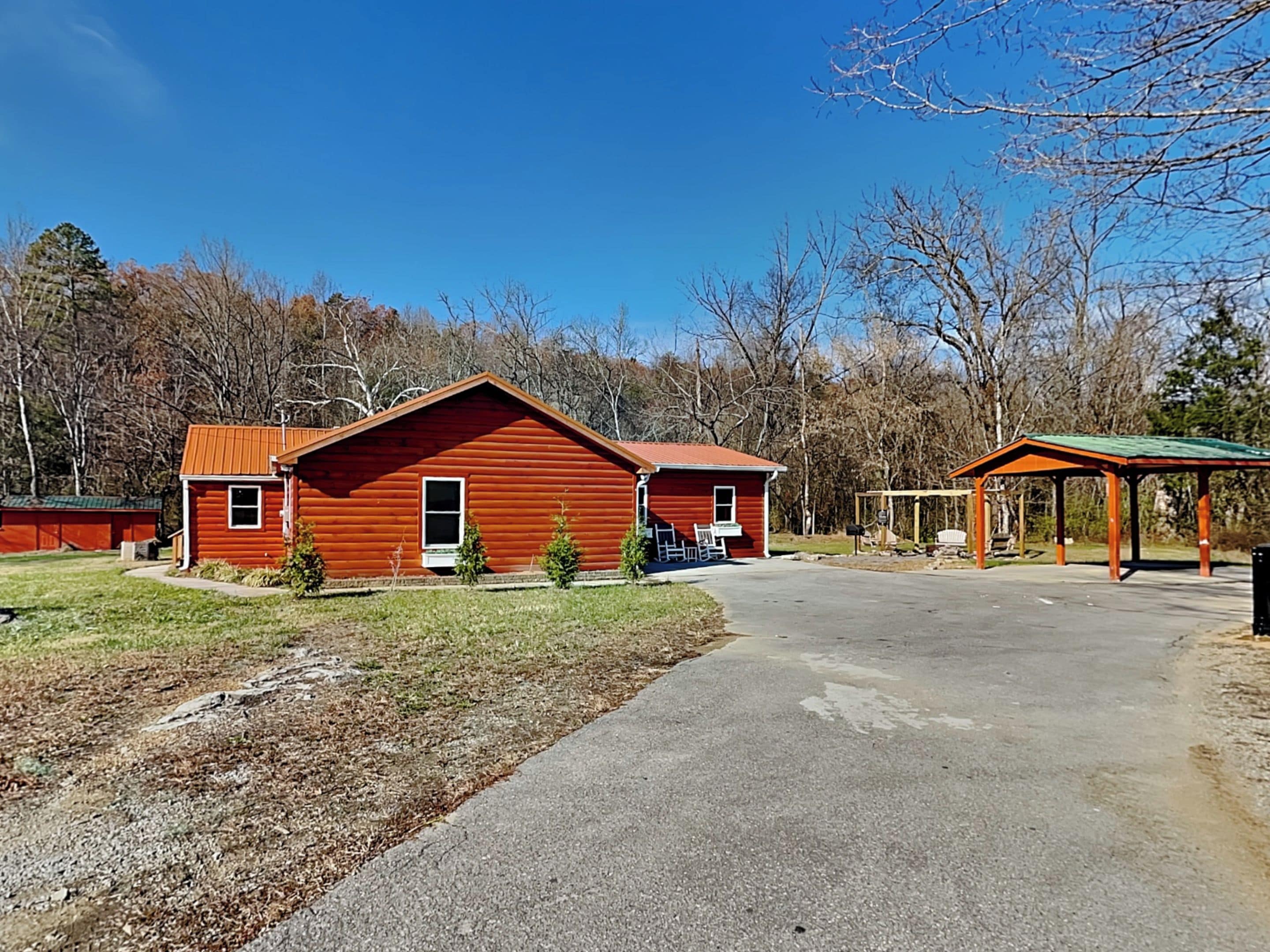A red wooden cabin with a green roof, a covered outdoor seating area, and a paved driveway.