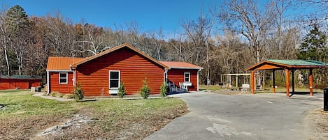 A red wooden cabin with a green roof, a covered outdoor seating area, and a paved driveway.