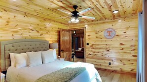 Une chambre aux murs lambrissés, avec un lit, un ventilateur de plafond et une porte donnant sur une autre pièce.