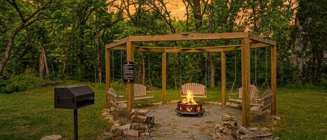 A wooden gazebo with a fire pit in the center, surrounded by chairs and a mailbox.