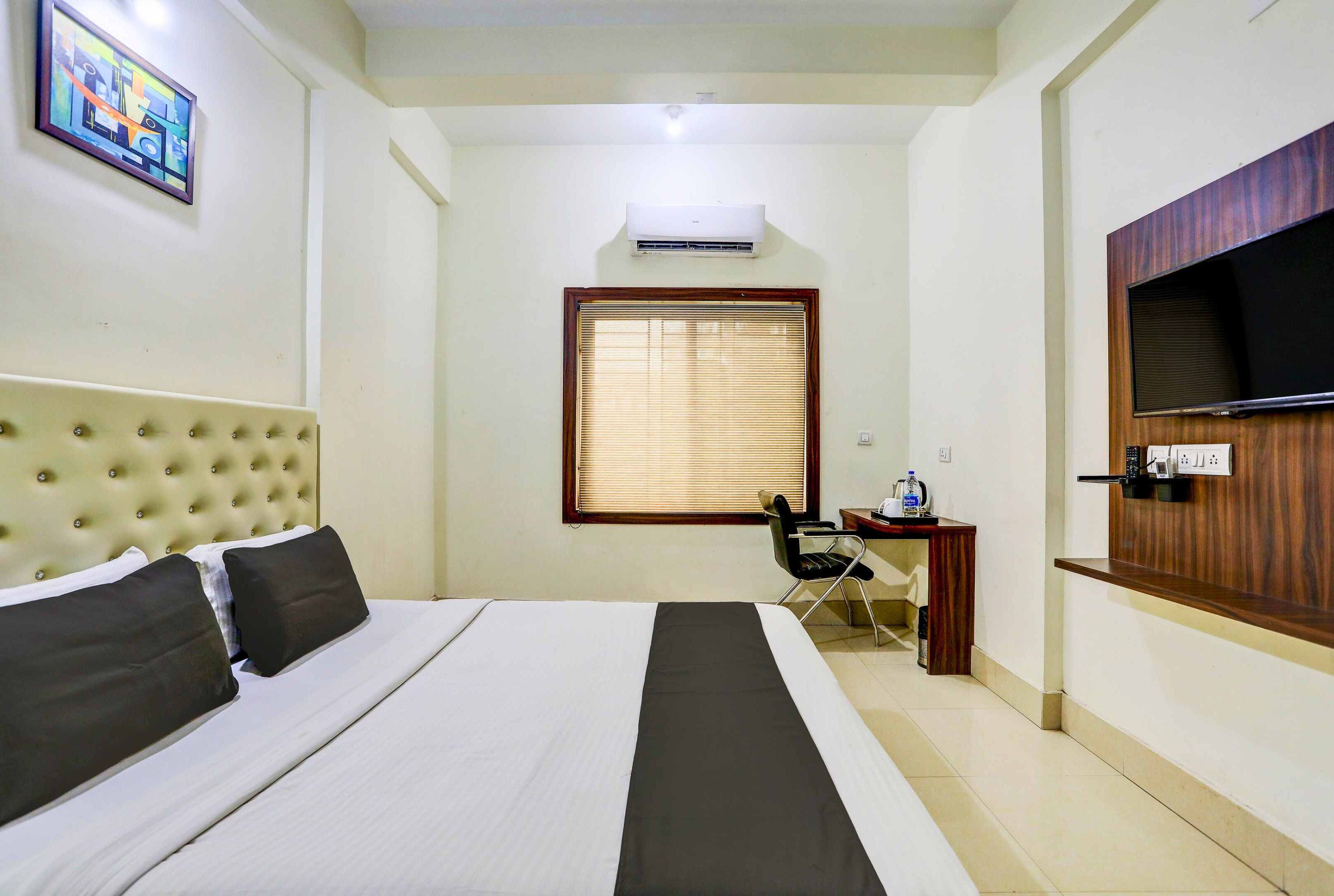 A hotel room with a bed, a desk, a chair, an air conditioning unit, a television, and a window with blinds.